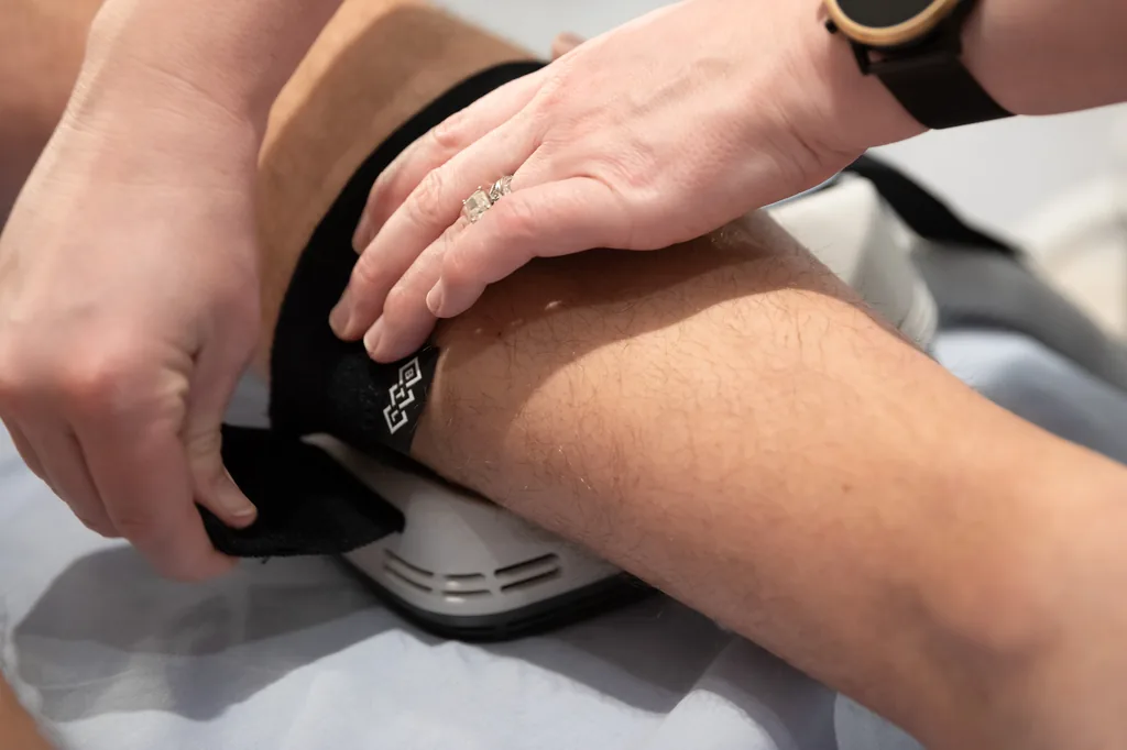 Hands fasten a black strap printed with a small diamond pattern around a client's calf, securing it to a white pad.