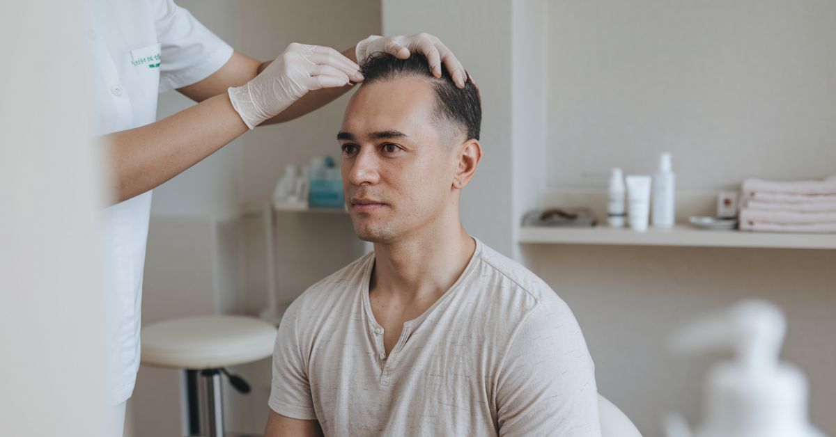 Gloved hand parting a seated man's hair to examine his scalp, shelves of towels and bottles in the treatment room behind.