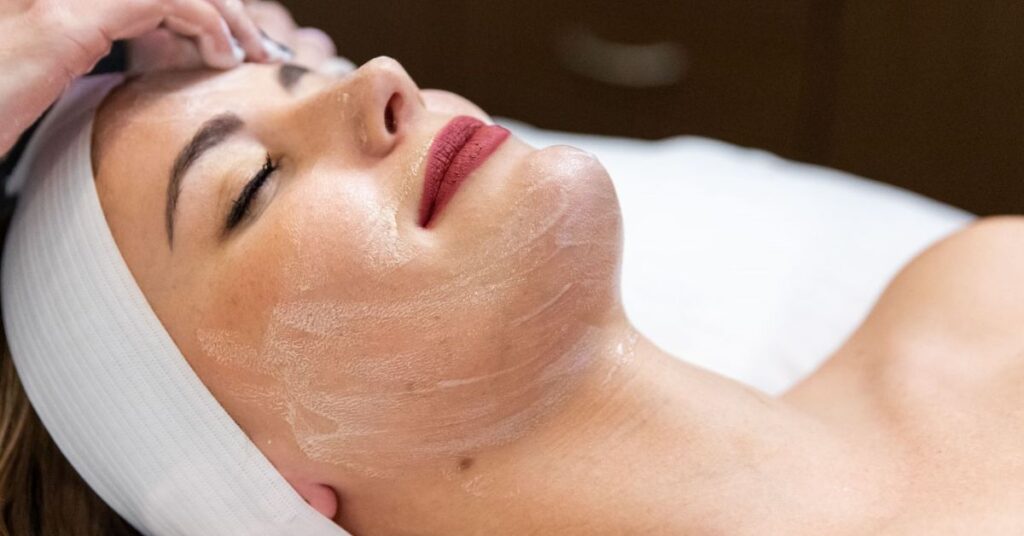 Woman lying down with eyes closed and a white cream mask applied to her cheek and jaw, glossy pink lips, white head wrap.