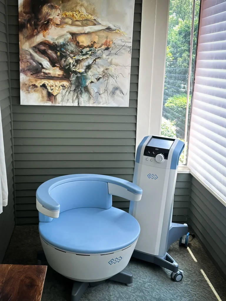 A rounded blue treatment chair beside a tall control console in a room with sage green paneled walls and an abstract painting.