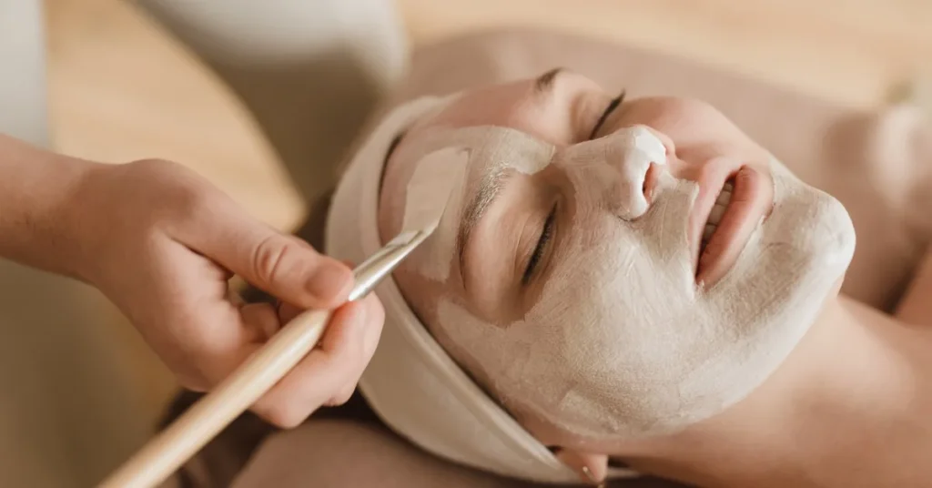 A hand uses a small brush to apply a light colored face mask to a smiling woman wearing a cloth headband.