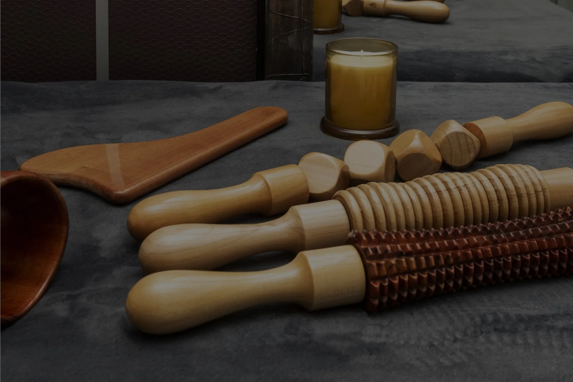 Carved wooden massage tools including rollers, a paddle, and a small bowl, arranged on a gray cloth with a lit candle behind them.