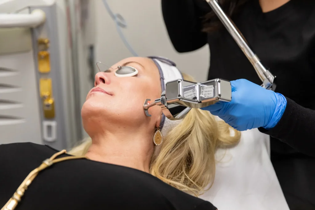 A gloved hand holds a chrome laser handpiece near a reclined client's cheek, who wears round metal eye shields and a headband.