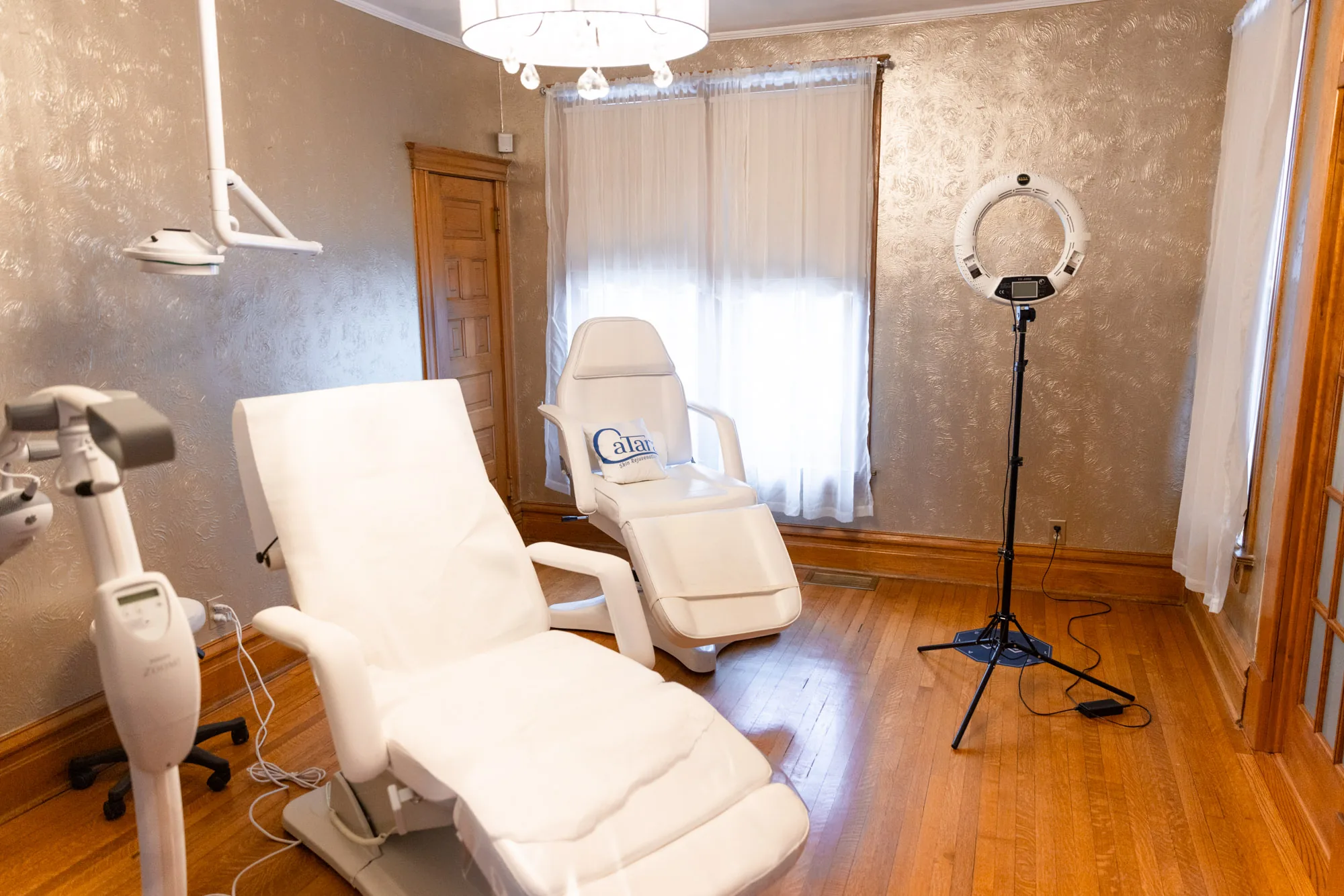 Empty treatment room with a reclined white spa chair, an articulated overhead light, and a ring light on a tripod stand.