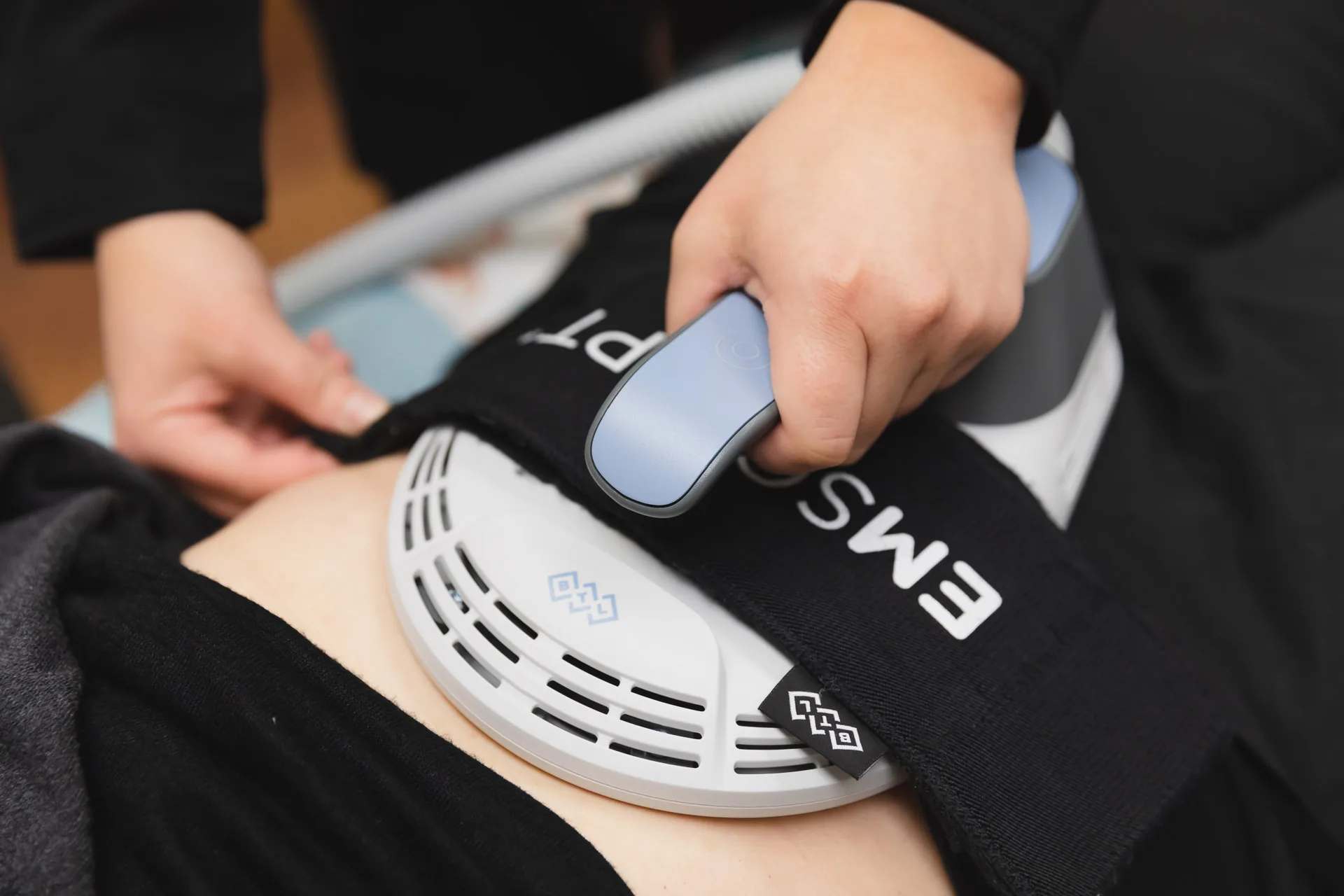 A hand holds a blue and black Emsculpt applicator secured by its strap against a person's bare abdomen.
