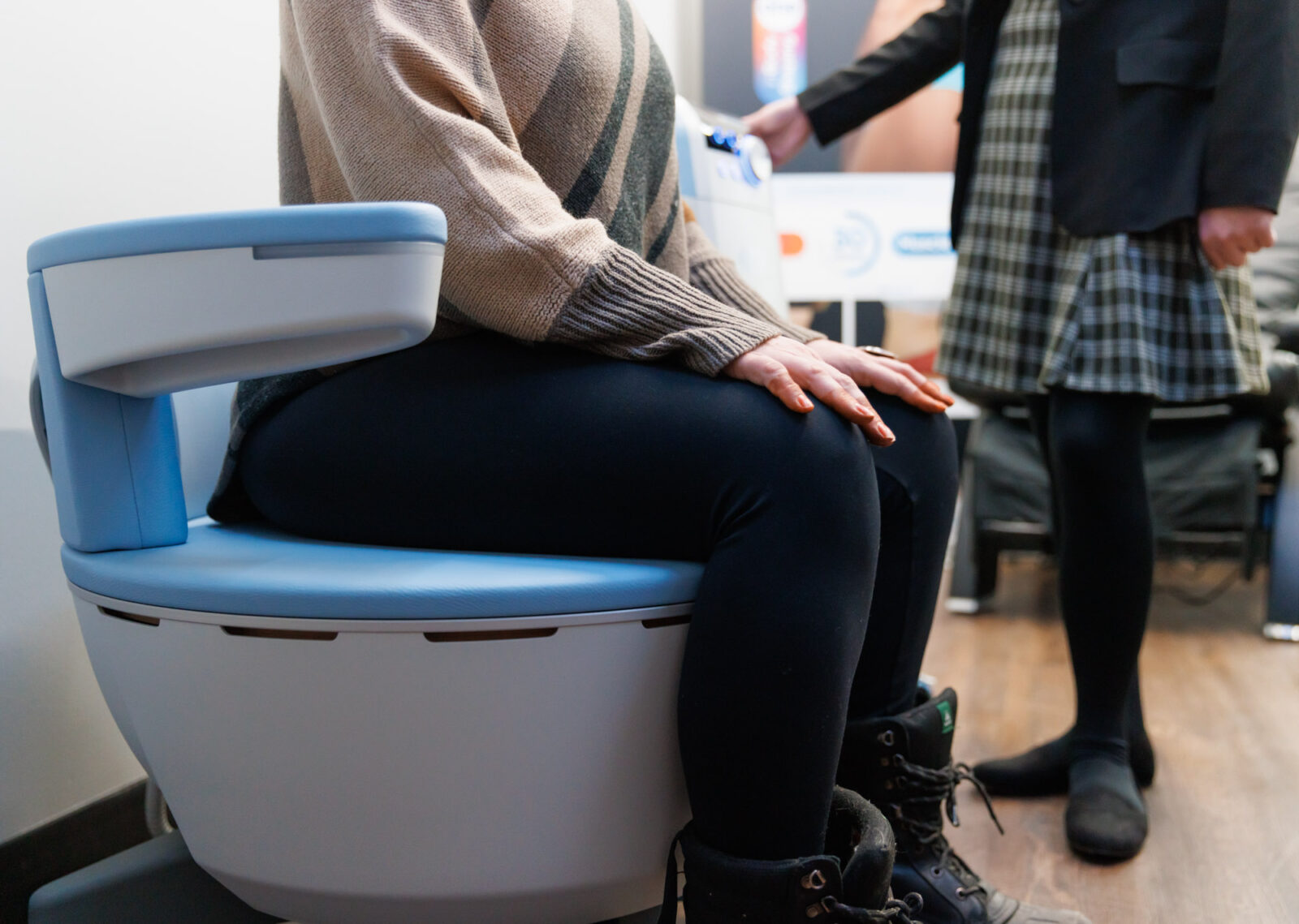 Person in a tan sweater seated on a blue cushioned treatment chair, hand on knee, clinician standing nearby holding a device.