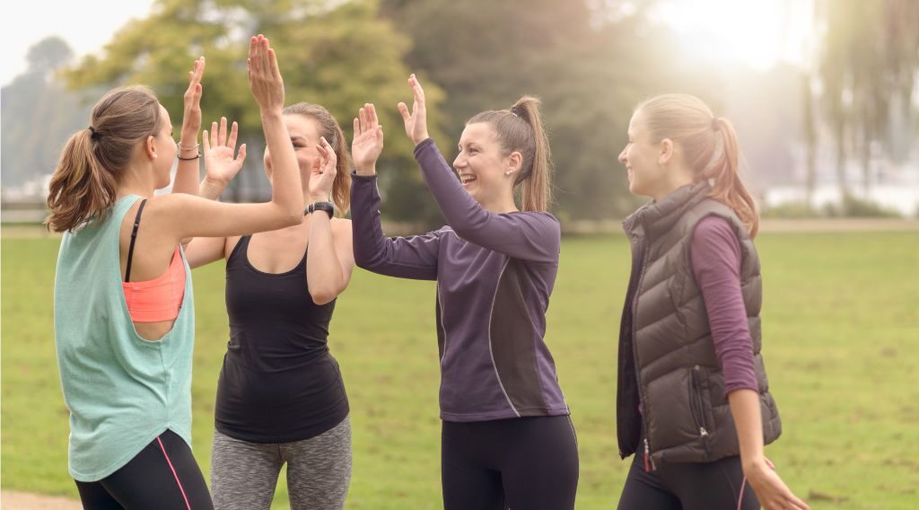 Four women in athletic wear high-fiving each other outdoors in a sunlit grassy park.
