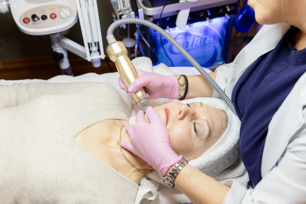 Gloved hand holds a gold-toned wand connected by a coiled cord against a reclining client's neck, an illuminated panel visible on equipment behind them.