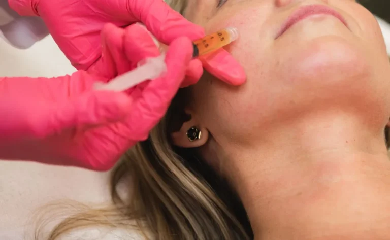 Gloved hand in pink injecting a syringe of amber liquid into the cheek of a person with a black stud earring.