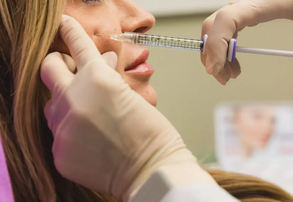 Gloved hand injects a marked syringe near a client's nasolabial fold and upper lip, a blurred photo behind.