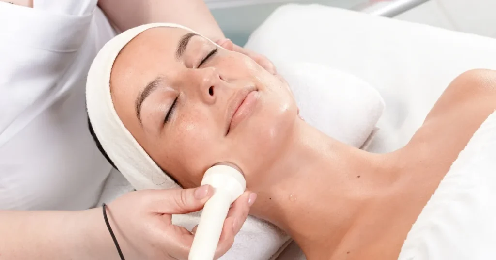 Clinician's hand holding a white handheld wand with a rounded tip against a client's jaw, eyes closed, white head wrap.