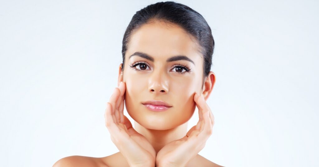 Portrait of a woman with hair pulled back cupping her own jawline with both hands against a plain white background.