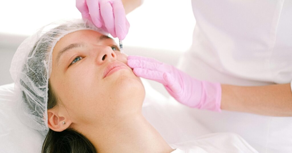 Pink gloved hands hold a fine metal instrument near the inner eyebrow and nose bridge of a reclining client in a mesh cap.