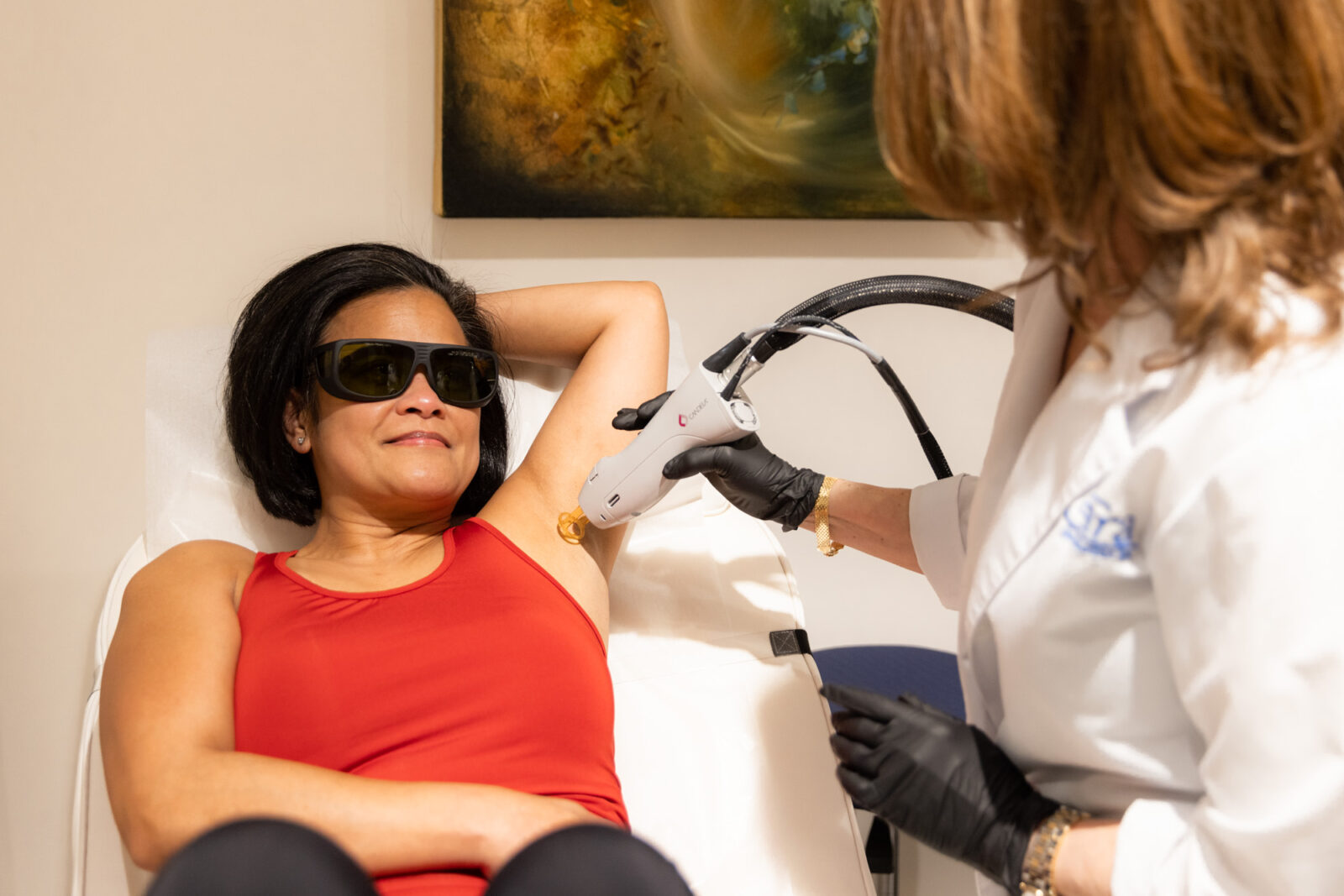 Clinician in a white coat holding a laser handpiece against a seated woman's underarm, a black hose attached.