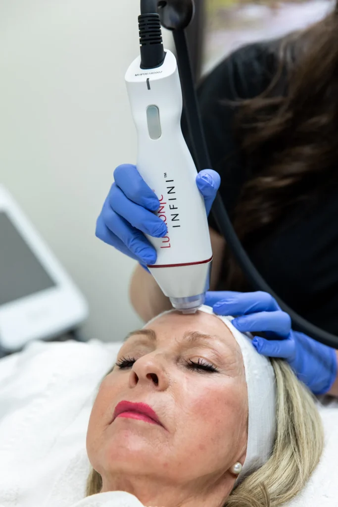 A gloved hand holds a Lutronic Infini handpiece against a reclining woman's forehead, her eyes closed and a white headband in her hair.