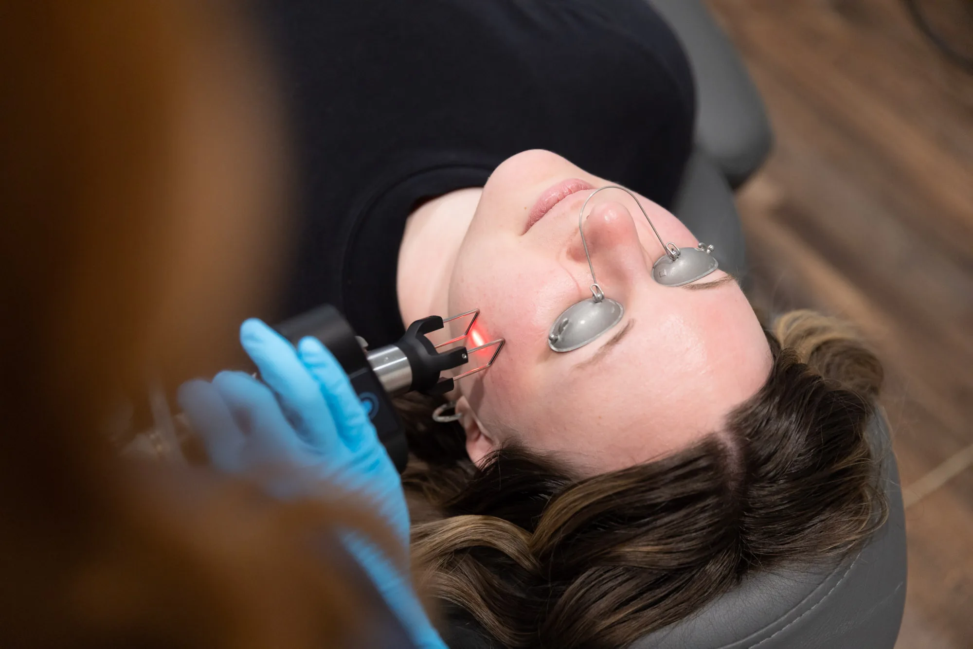 Gloved hand guiding a laser handpiece with a visible red aiming beam across a client's cheek during a laser facial at Catara Med Spa in Algonquin, IL, the client wearing protective eye shields.
