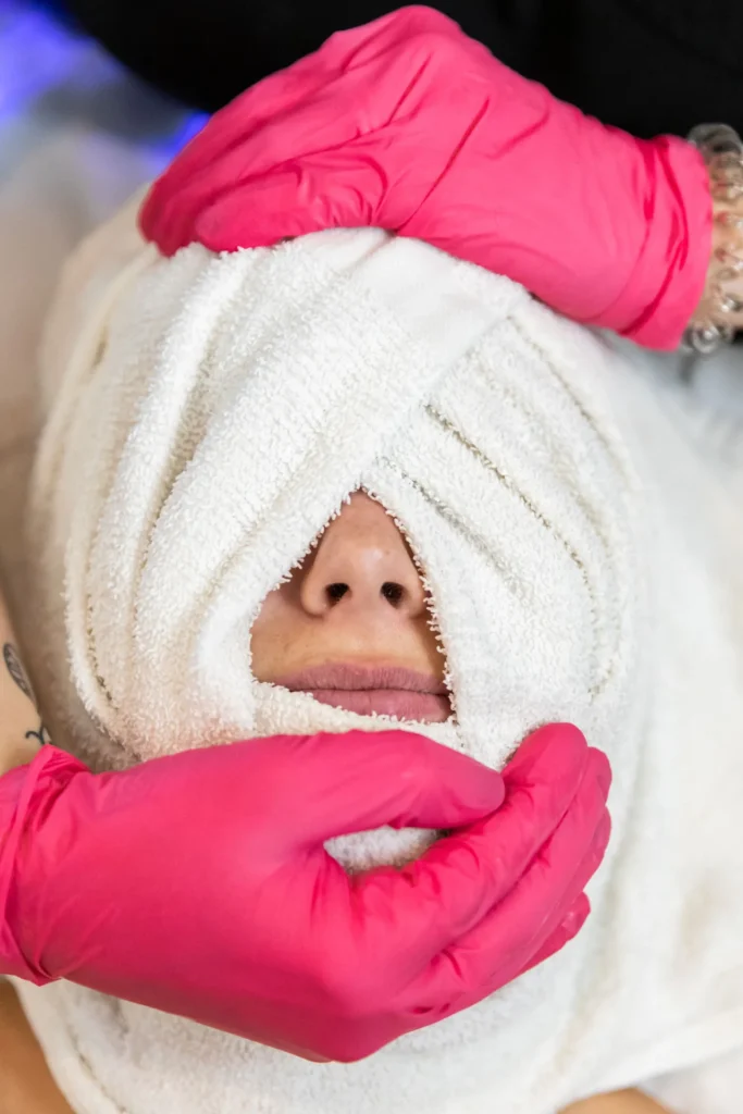 Pink-gloved hands wrap a white towel around a person's face, leaving only the nose and mouth visible.