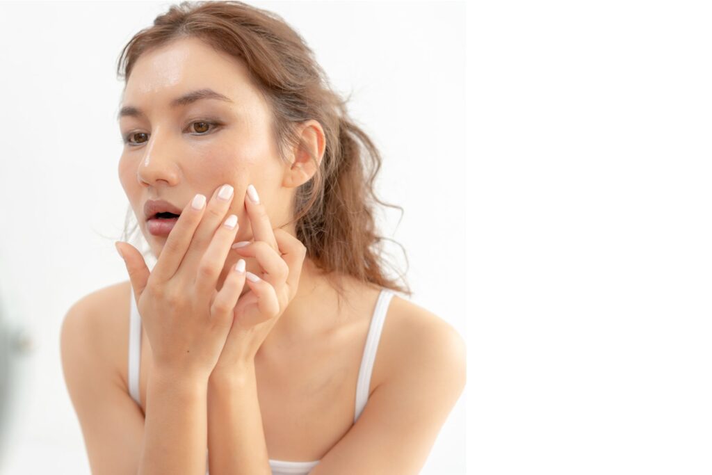A woman with wavy light brown hair touches her cheek with her fingertips, wearing an off white top against a softly blurred background.