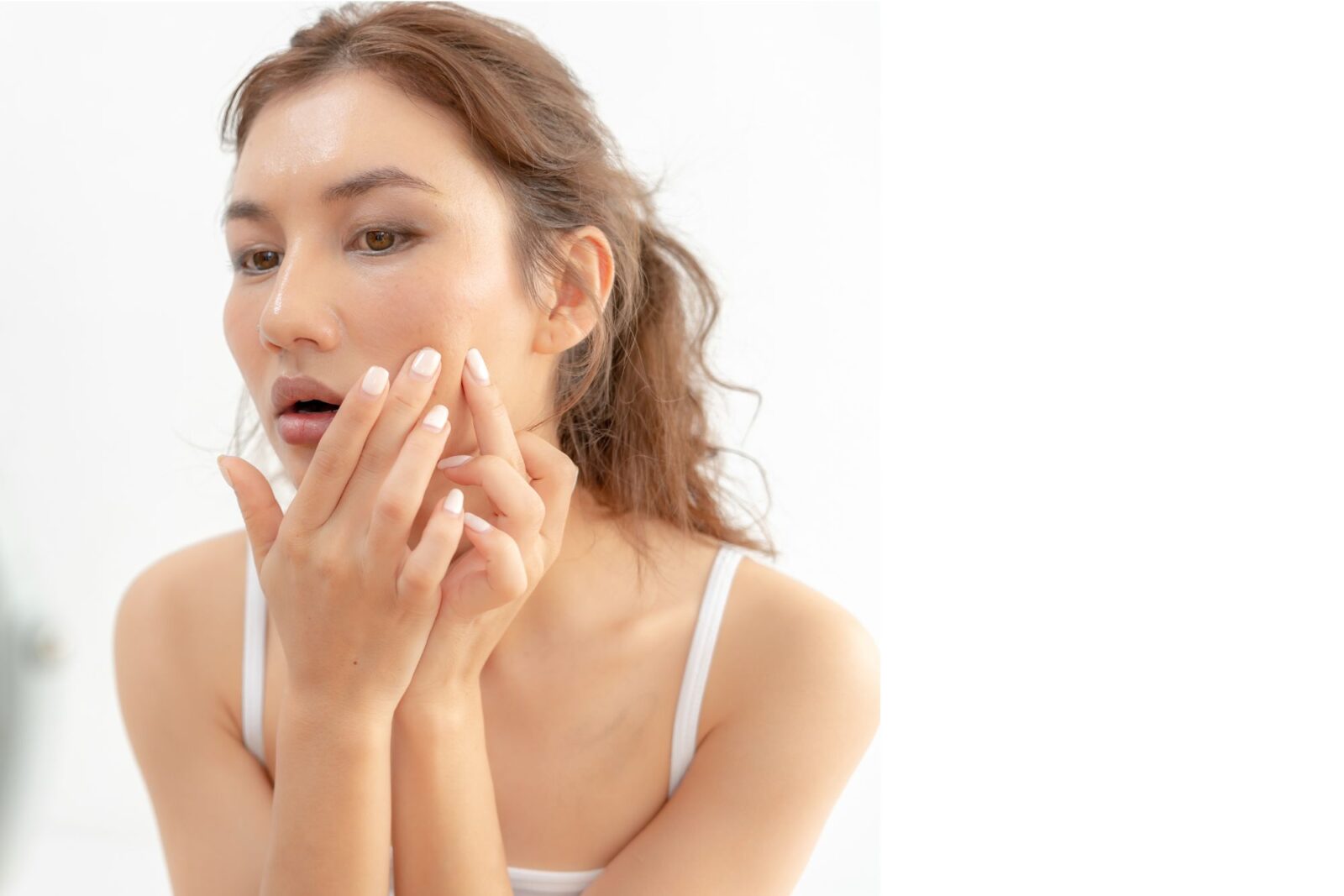 A woman with wavy light brown hair touches her cheek with her fingertips, wearing an off white top against a softly blurred background.