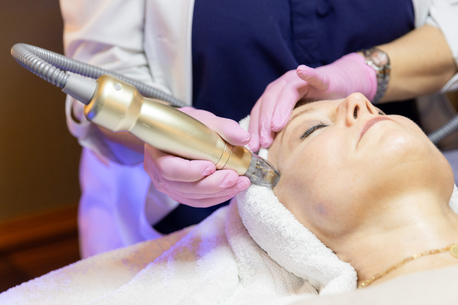 Gloved hand holds a gold-toned wand against a reclining client's temple, connected by a cord, blue-lit device visible behind them.