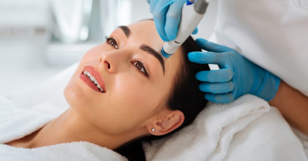 Woman reclined in white towels smiling while a blue gloved clinician holds a handheld skincare device near her forehead.