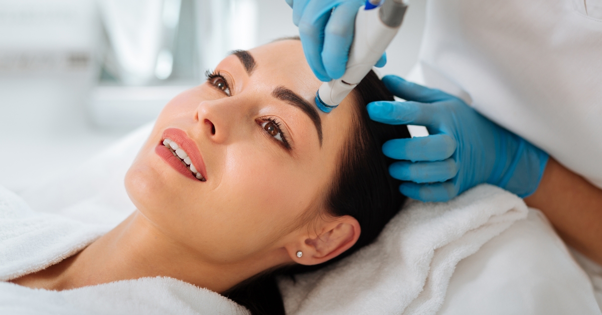 Woman reclined in white towels smiling while a blue gloved clinician holds a handheld skincare device near her forehead.