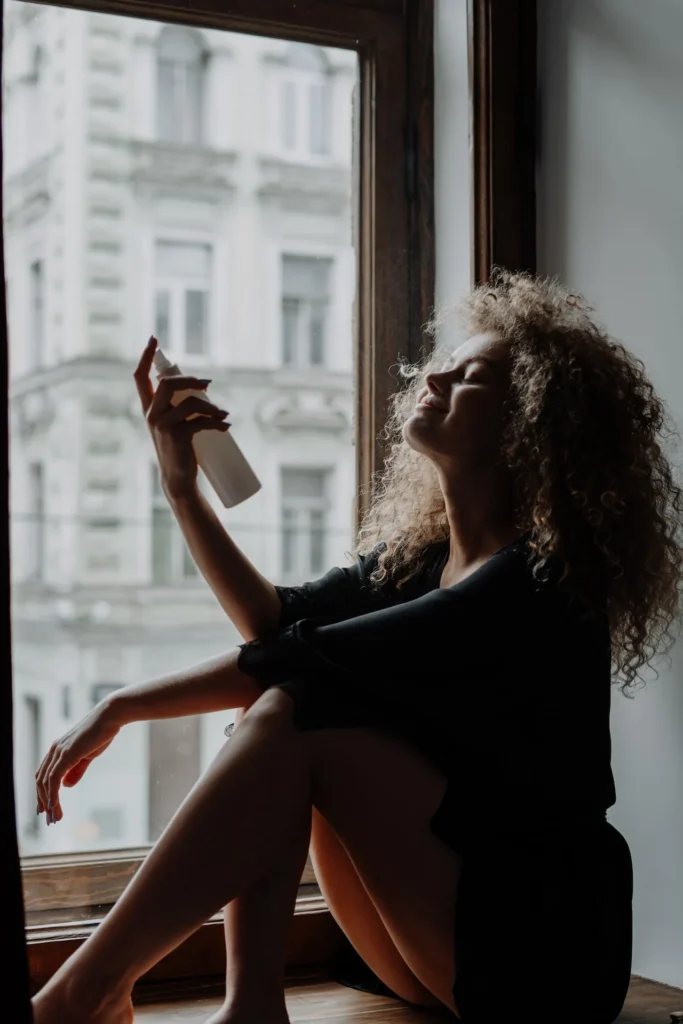 A woman in silhouette sits on a windowsill, holding a white spray bottle near her face, eyes closed, city buildings outside the window.