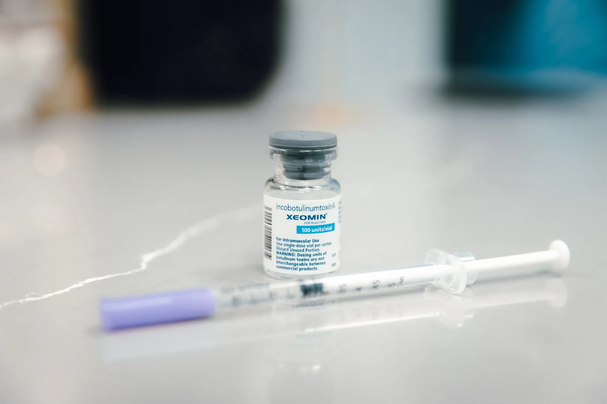 A Xeomin incobotulinumtoxinA 100 units per vial bottle on a reflective counter next to a syringe with a lavender cap.