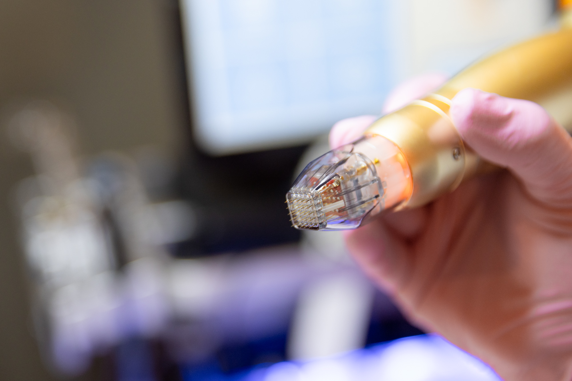 Close-up of a gold-colored handpiece held in a gloved hand, fitted with a clear cartridge tip holding a grid of fine needles.