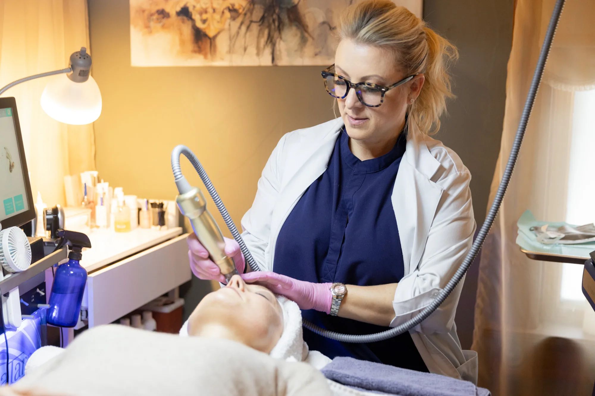 Clinician in a white coat guiding a hose-connected hydradermabrasion wand across the face of a reclining client during the HydraFacial step of the three week Glass Skin Facial series at Catara Med Spa in Algonquin, IL