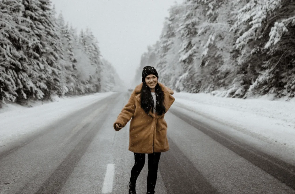 Woman in a camel coat and black beanie walking toward the camera on a snow-covered road lined with snowy evergreen trees.
