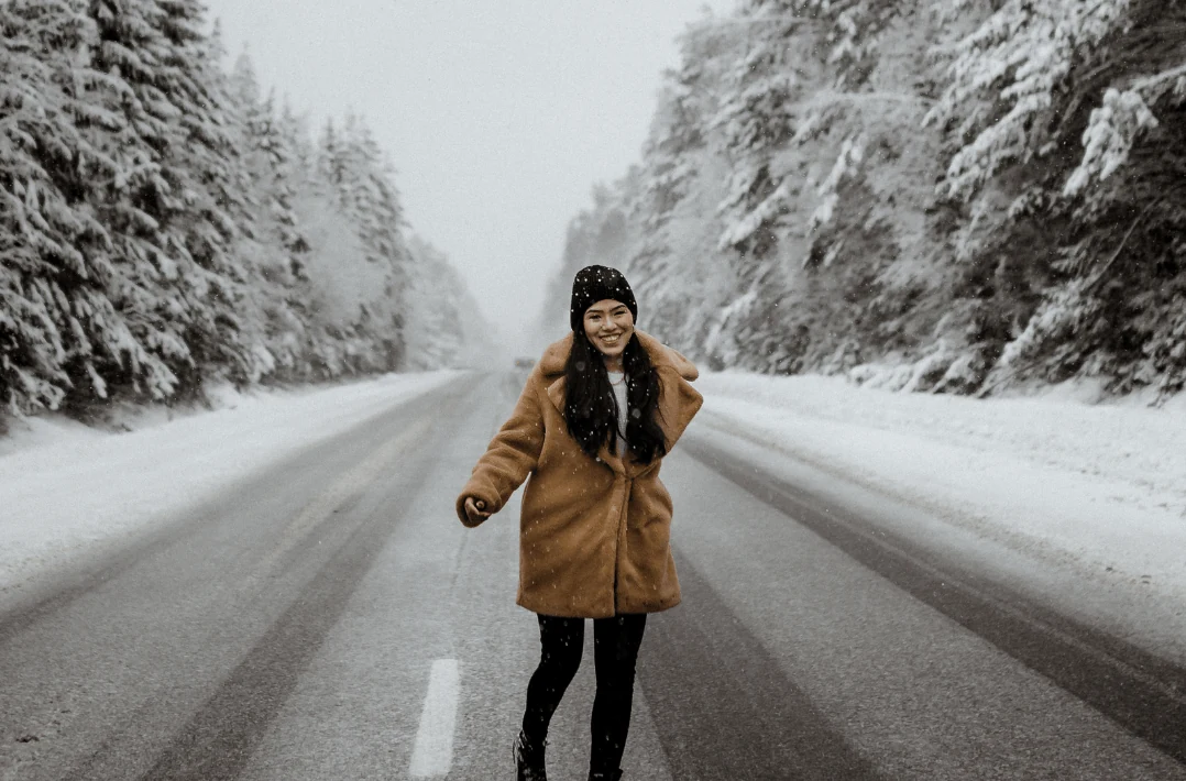 Woman in a camel coat and black beanie walking toward the camera on a snow-covered road lined with snowy evergreen trees.
