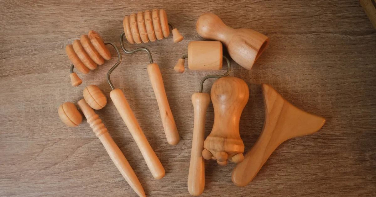 An assortment of wooden massage tools, including ridged rollers and curved handled pieces, laid out on a wood grain table.